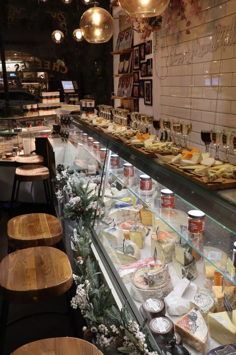 The counter at Cheeses of Muswell Hill laid for a Cheese Lates evening, with boards of cheese, rows of wine glasses and wooden stools
