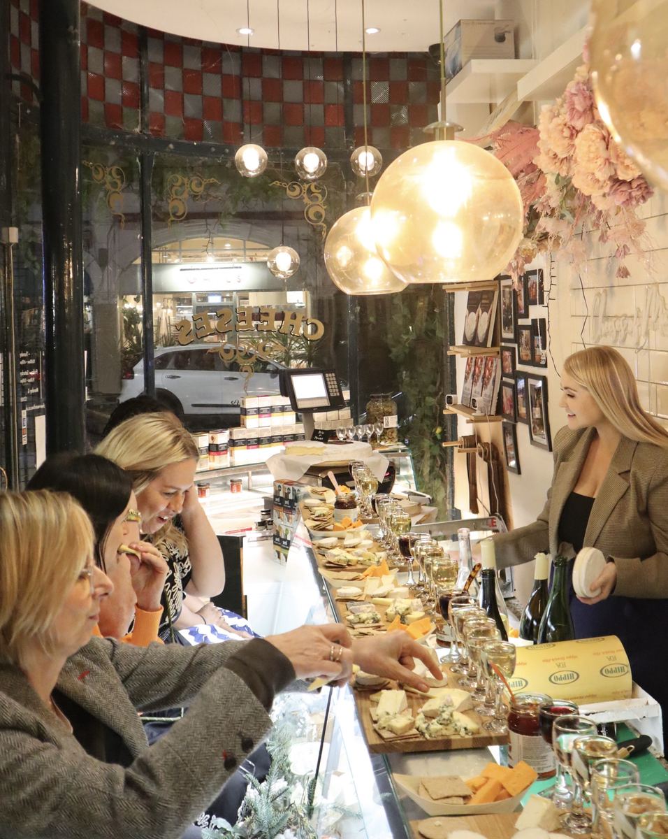 Morgan McGlynn Carr serves guests at a Cheese Lates tasting inside Cheeses of Muswell Hill, with boards of cheese and glasses of wine along the counter