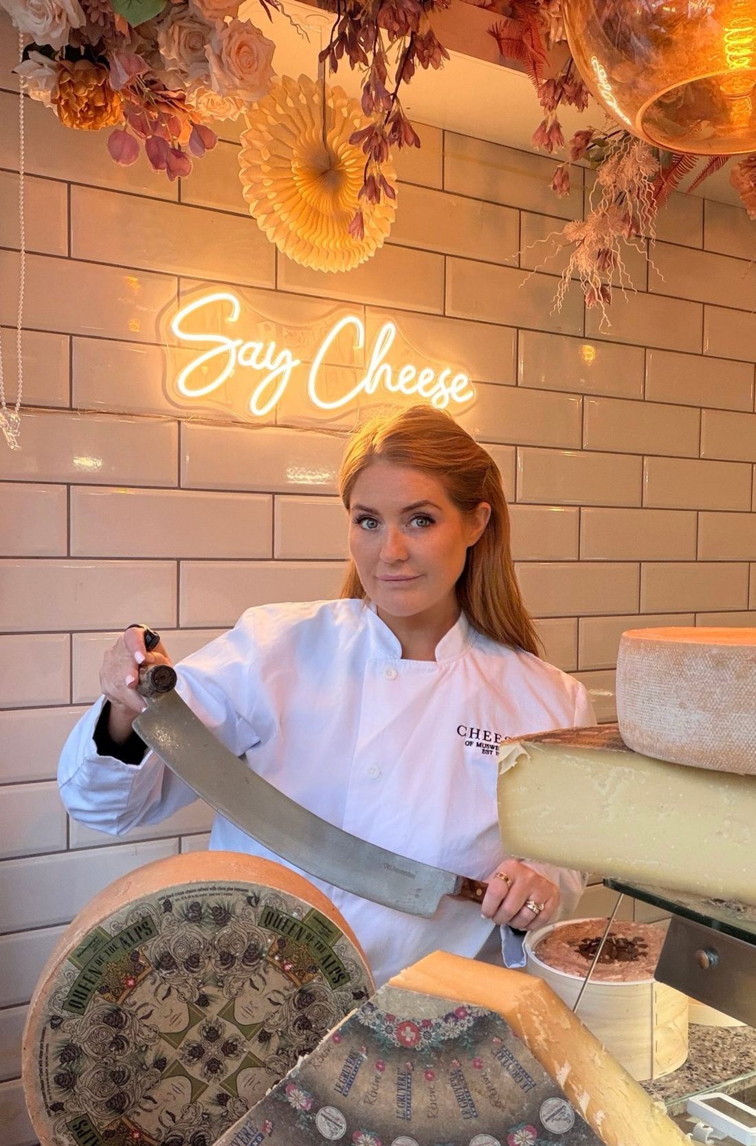 Morgan McGlynn Carr, UK cheese expert and cheesemonger, holding a cheese knife beside wheels of Alpine cheese at Cheeses of Muswell Hill, London
